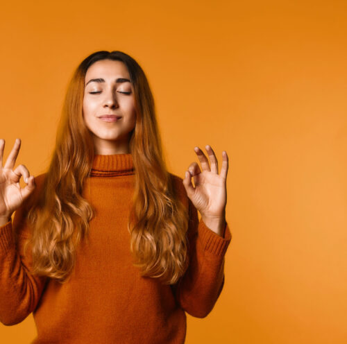 Calm redhead caucasian girl dressed in sweater of ocher color on the orange background, with closed eyes, is concentrating and relaxing